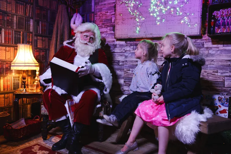 Father Christmas reading from his book to two young children in the Cabin
